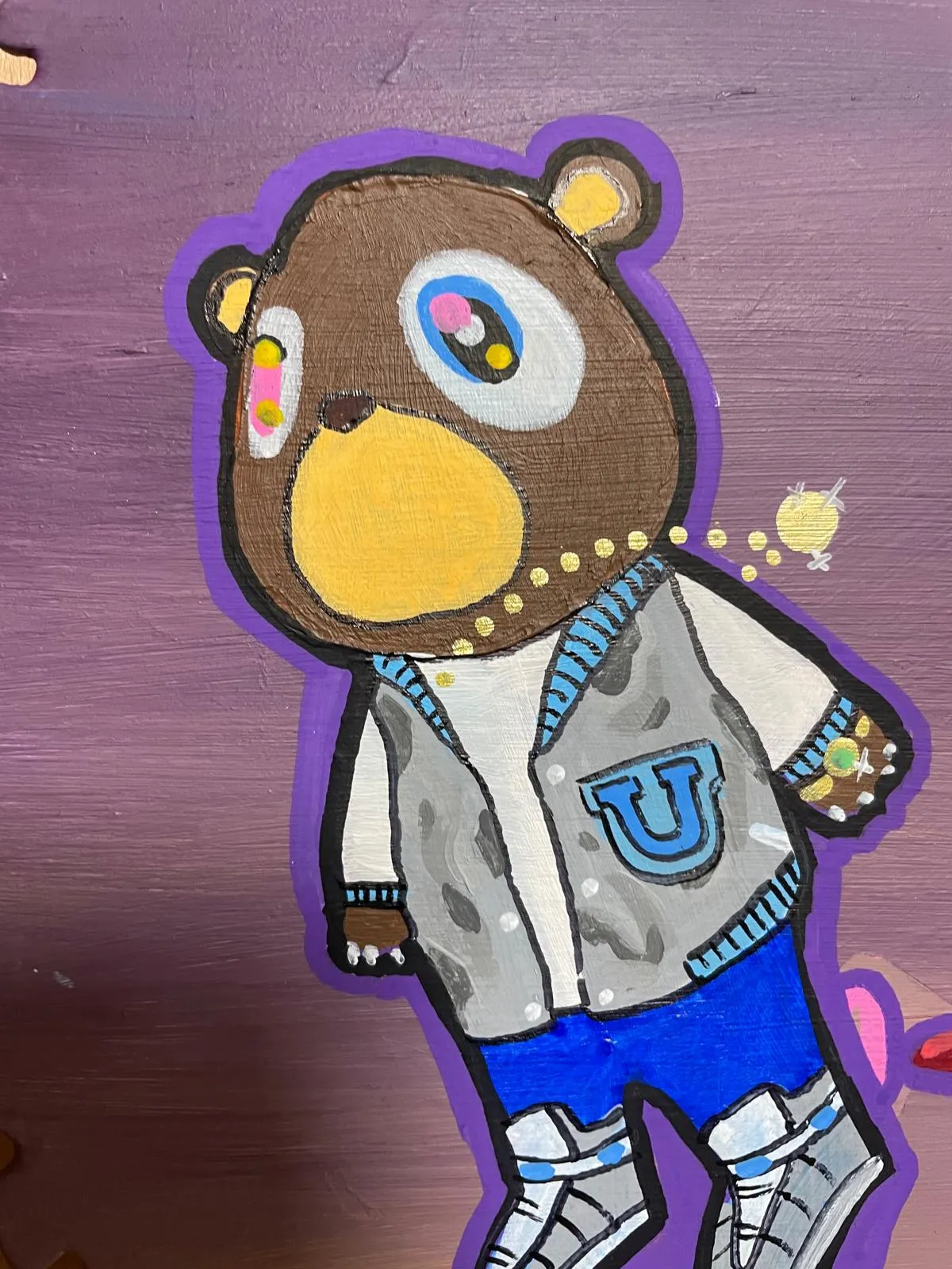 Close view of the bear on the blade: a brown bear with a mismatched eye and a yellow muzzle, wearing a grey varsity jacket with a blue U on the chest and blue shorts, outlined in purple against a purple ground.