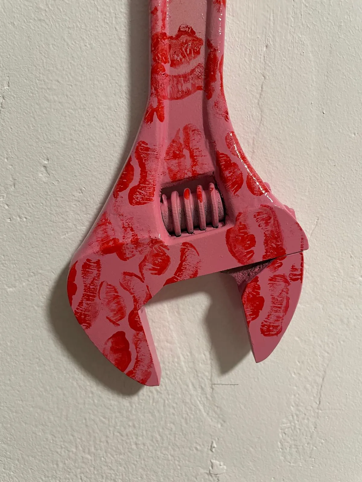 The pink wrench on a white wall, cropped to the adjustable jaw and worm screw, crowded with red lipstick prints.
