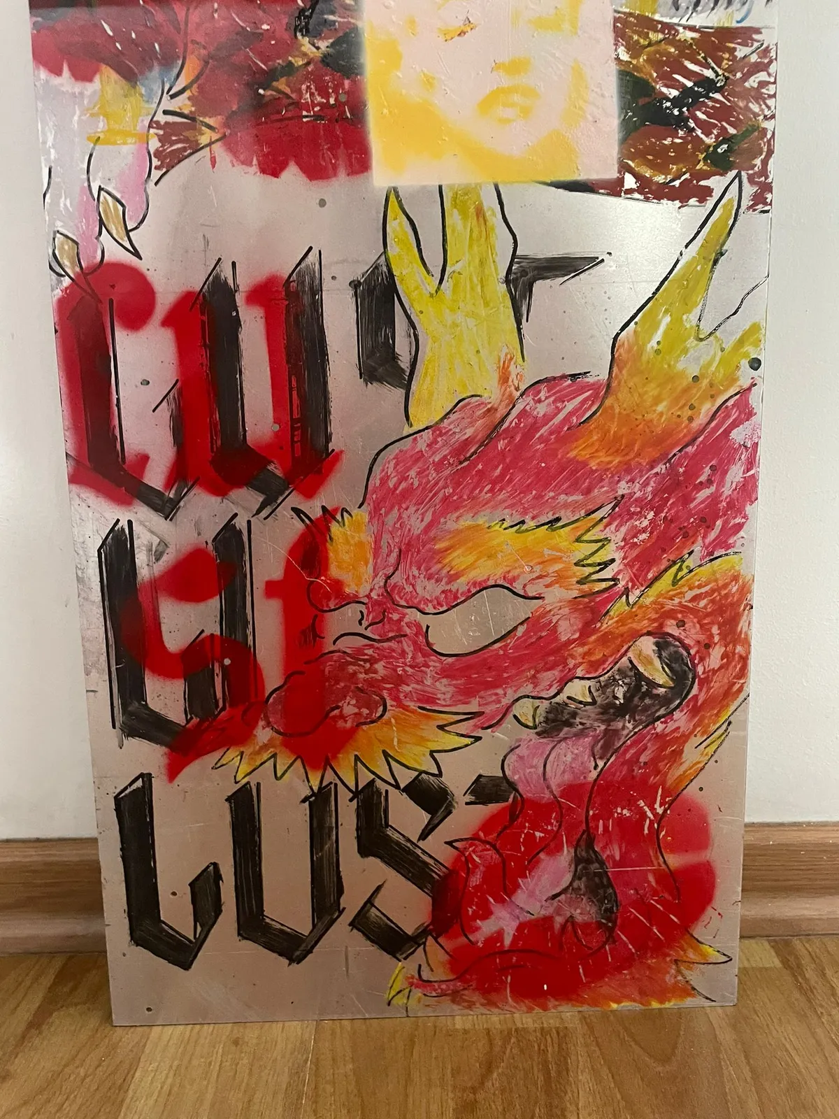 The metal panel standing on a wooden floor against a white wall. The blackletter word LUST is stacked down the left; a phoenix in red, orange and yellow oil pastel flies across the right.