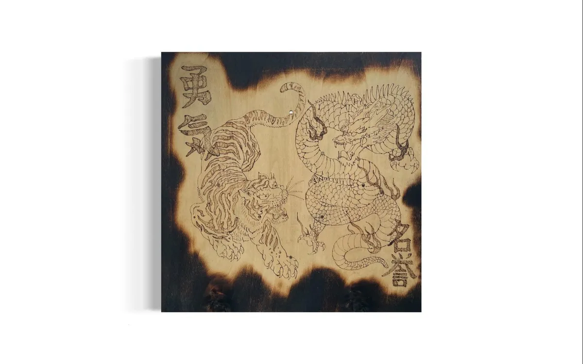 A square plywood panel with a tiger and a coiled dragon burned into the pale grain in fine line, columns of kanji at the upper left and lower right, and a heavily scorched black border around all four edges.
