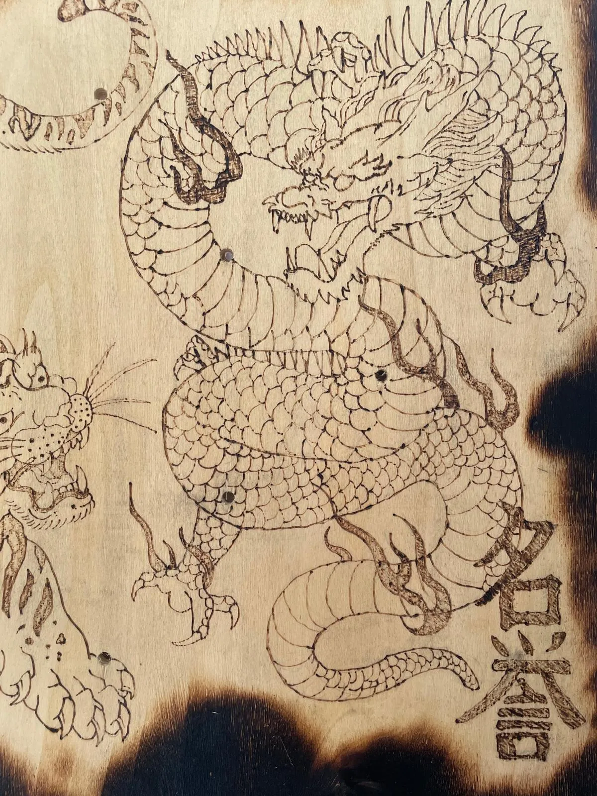 Detail of the burned dragon: scaled body, clawed feet and whiskered head drawn in thin scorched line, with a column of kanji burned beside it.